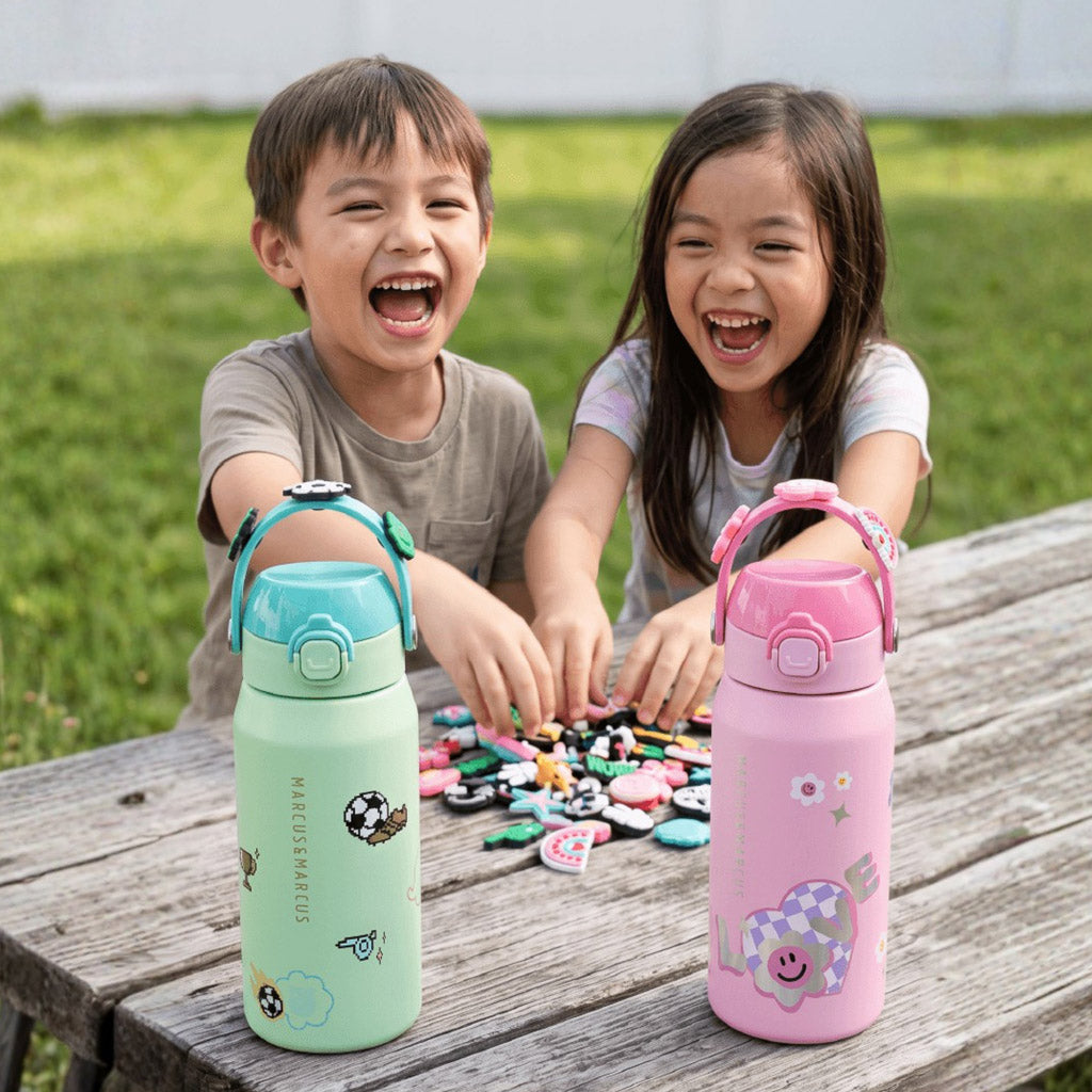Two smiling children customizing their Marcus & Marcus CharmPoP insulated water bottles with charms outdoors